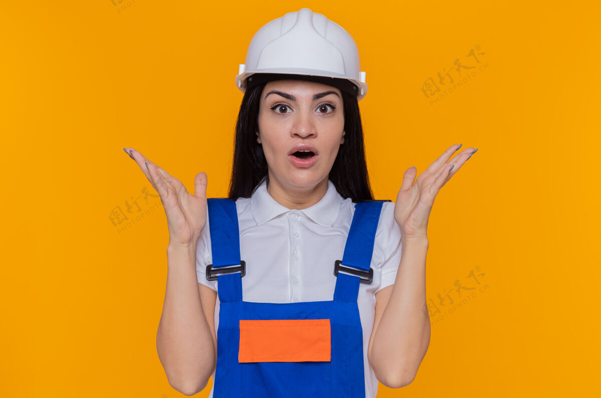 头盔身穿施工制服 头戴安全帽的年轻建筑妇女站在橘色的墙上 一边高兴一边惊讶地举起双臂看着前方快乐站立安全