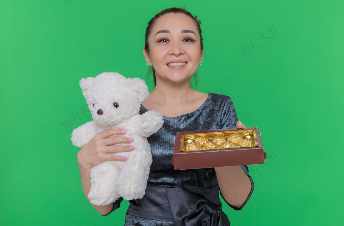 国际快乐的亚洲女人拿着泰迪熊和一盒巧克力糖果作为礼物微笑着站在绿色的墙上欢庆国际妇女节快乐亚洲盒子