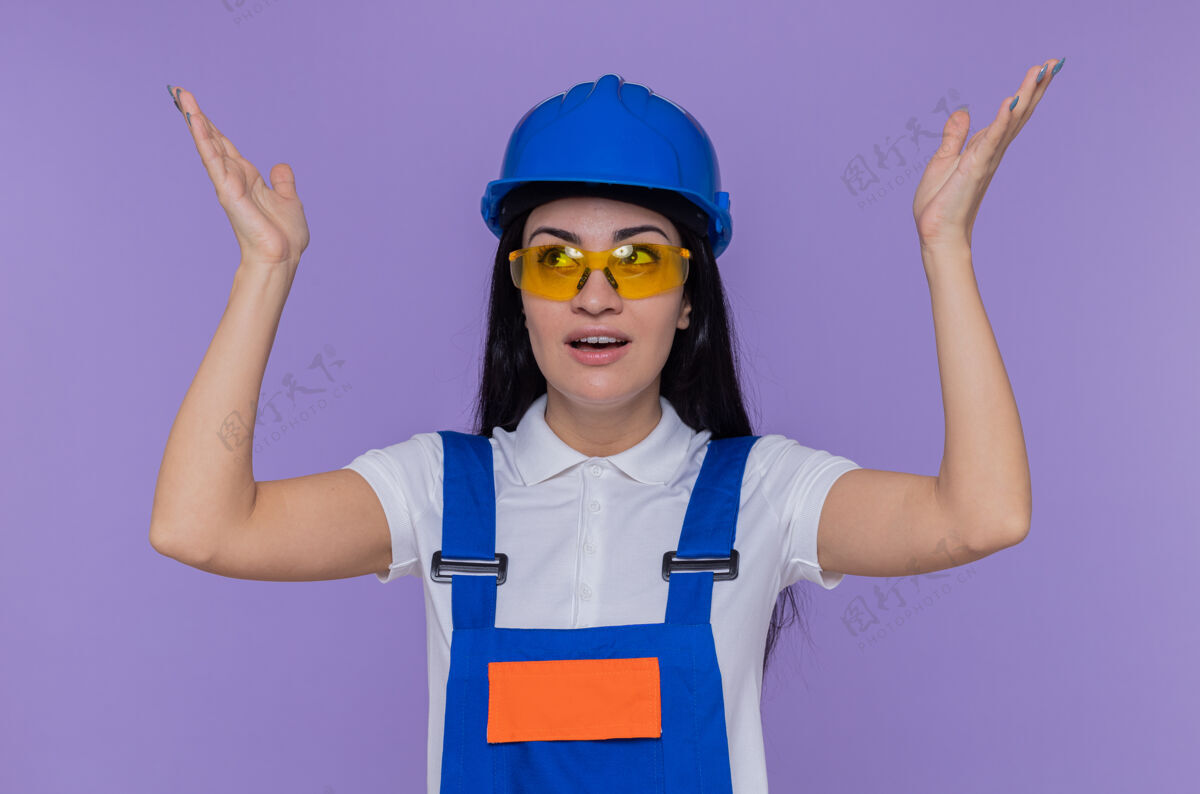 手臂身穿施工制服 头戴安全帽的年轻建筑女工人抬起双臂站在紫色的墙壁上 既高兴又惊讶安全惊喜女人