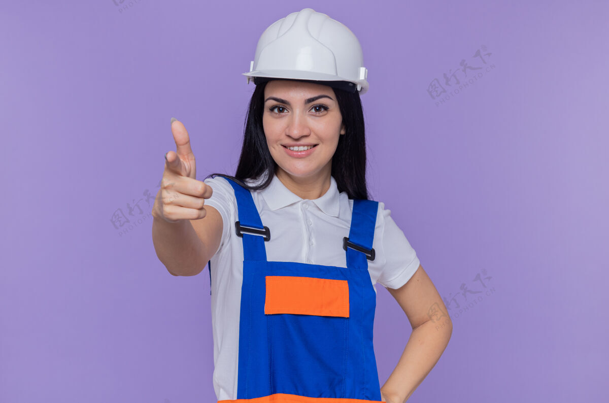指点身穿施工制服 头戴安全帽的年轻建筑工人自信地微笑着 用食指指着前面站在紫色的墙上安全女人站立