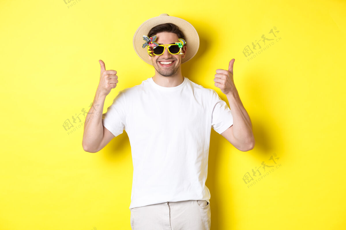 帅气旅游和生活方式的概念微笑的游客竖起大拇指 享受旅行和推荐 戴着夏季帽子和太阳镜 黄色背景的形象自信男人夏天