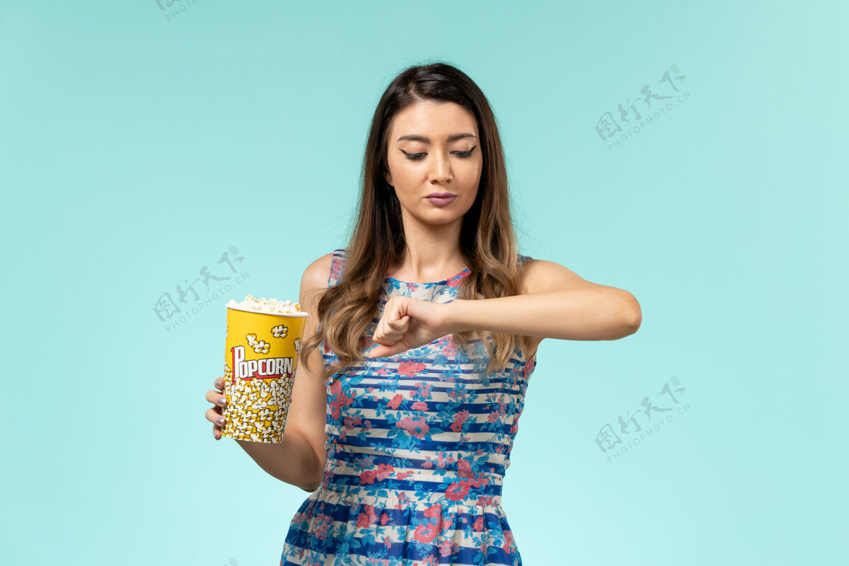 爆米花前视图年轻的女性拿着爆米花包 看着她的手腕在蓝色的表面剧院电影包装