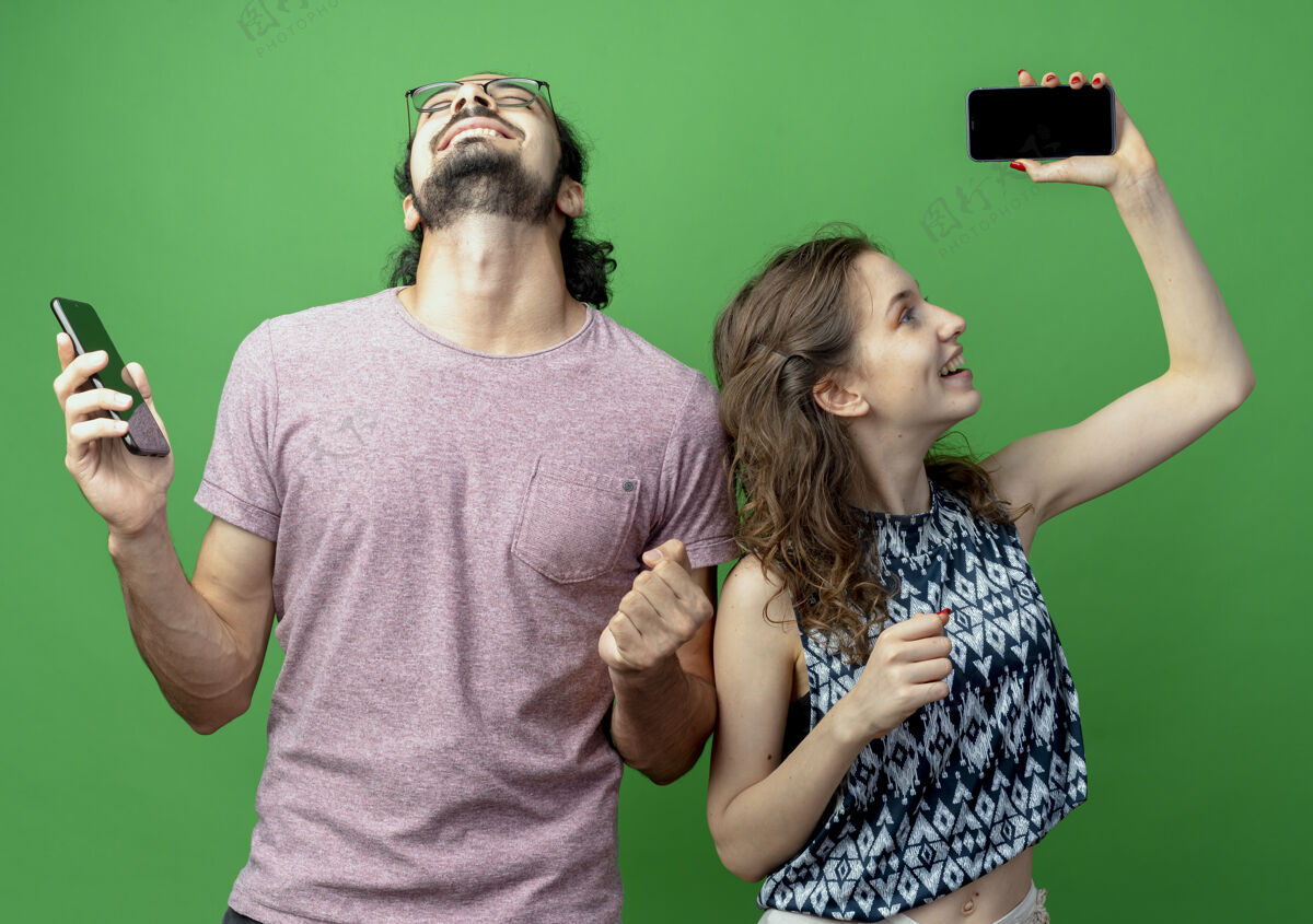 抱着一对年轻的夫妇站在绿色的墙边 高兴而兴奋地握着他们的智能手机握紧拳头男人年轻站着