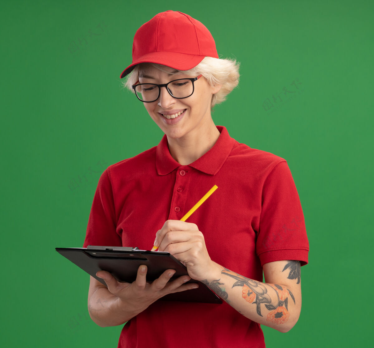 年轻年轻的送货员身穿红色制服 戴着帽子 戴着一副眼镜 上面有空白页的剪贴板和铅笔 站在绿色的墙上 脸上带着微笑写着什么微笑页写字