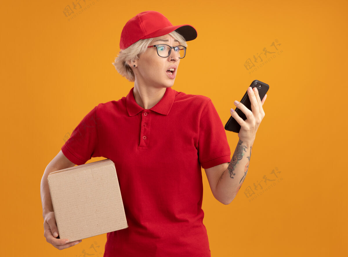 抱着身穿红色制服 戴着帽子 戴着眼镜 拿着纸板箱 看着智能手机的年轻送货员站在橙色的墙上 心神不安制服女人帽子