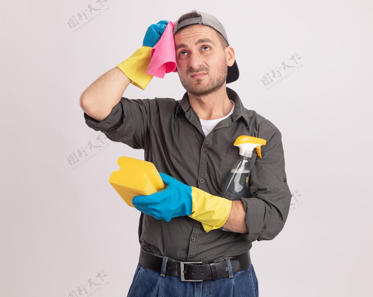 破布年轻的清洁工穿着便服 戴着橡胶手套 戴着帽子 肩上扛着清洁喷雾和抹布的海绵 站在橘色的墙上 困惑地抬起头来穿男人肩膀