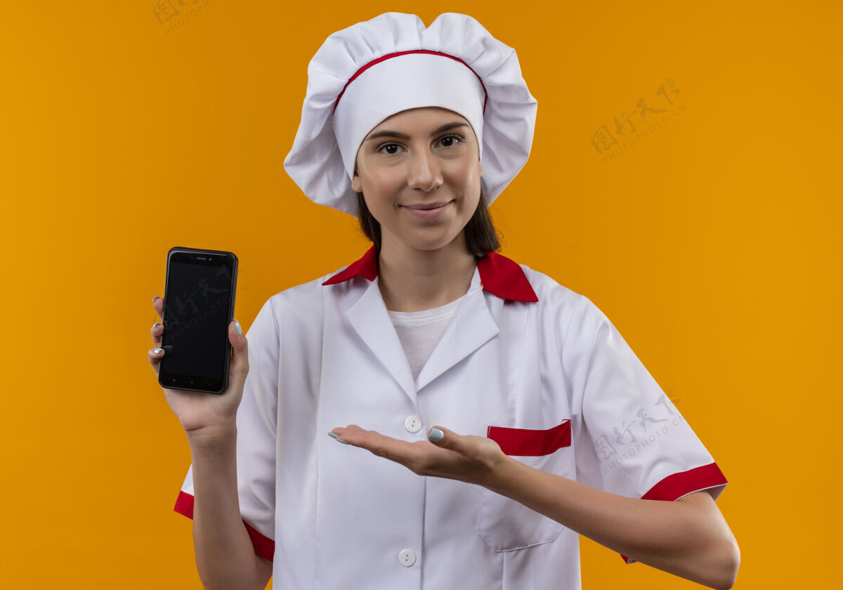 请身着厨师制服的年轻高加索厨师女孩高兴地拿着电话 指着橙色空间里的电话 还有复印空间点厨师制服