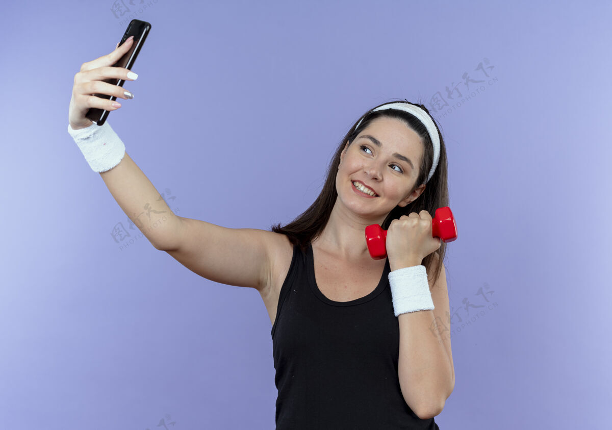 脸戴着头巾的年轻健身女用智能手机自拍手持哑铃微笑着幸福的脸站在蓝色的墙上移动使用拿着