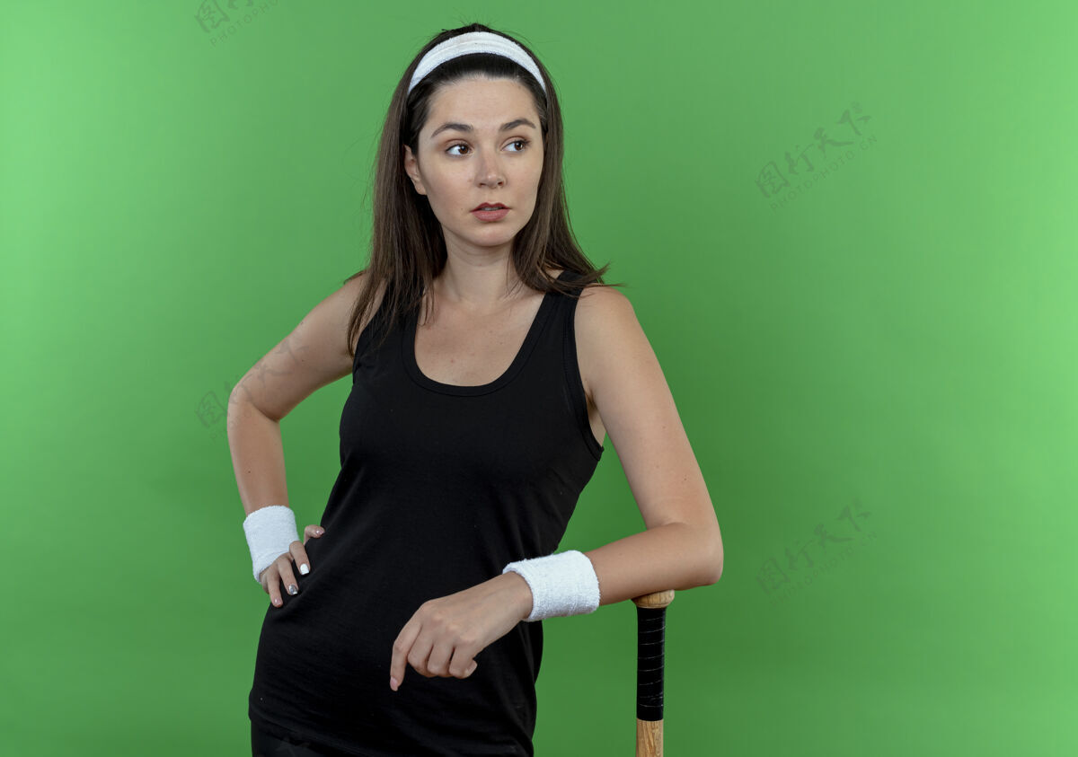 站立戴着头巾的年轻健身女 手靠在棒球棒上 自信地站在绿色的墙上 望向一边头带靠边年轻