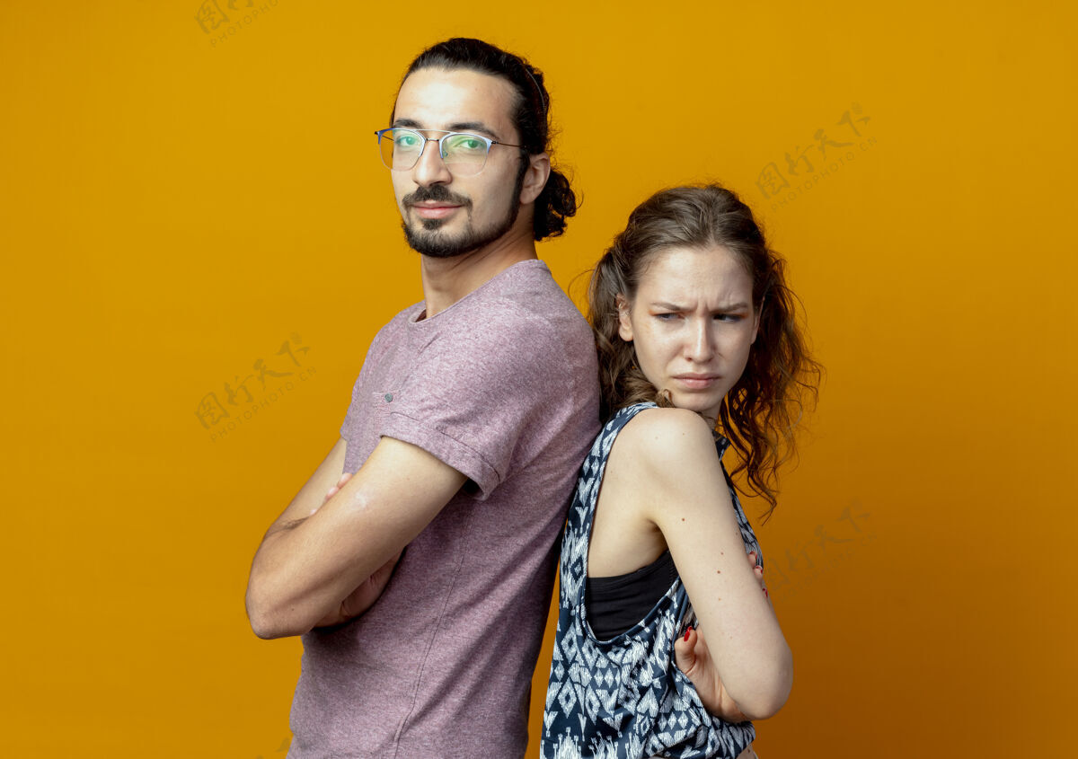 背这对年轻的夫妇背靠背地站在橙色的墙上 双臂交叉一对站手臂