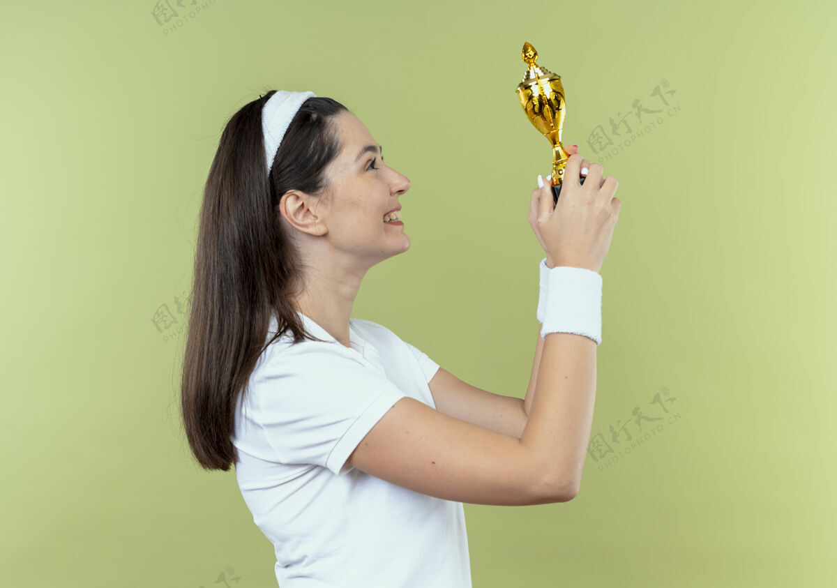 年轻戴着头巾的年轻健身女士拿着她的奖杯 高兴而兴奋地看着它站在轻墙上奖杯站着头带