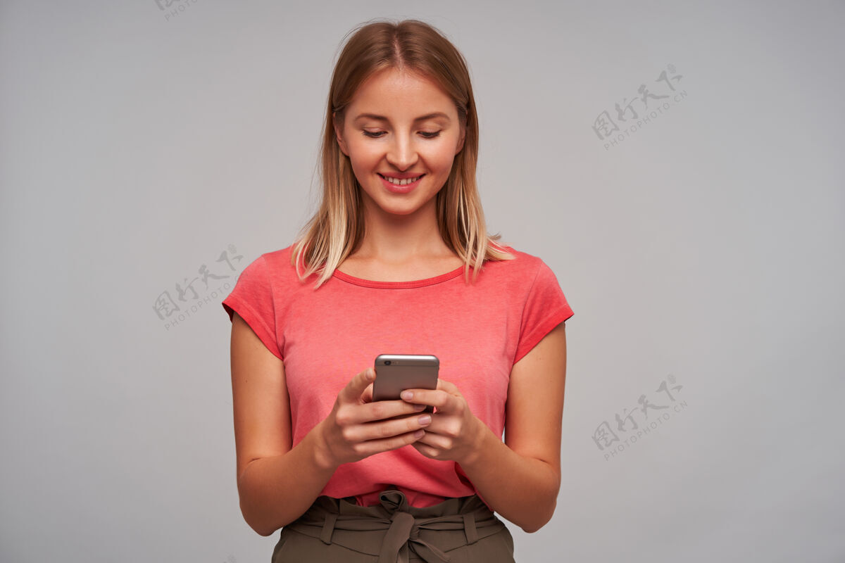 在线漂亮的女人 漂亮的金发女孩穿着粉色t恤和棕色裙子拿着智能手机微笑着看在灰色的墙上读朋友的留言皮肤漂亮年轻