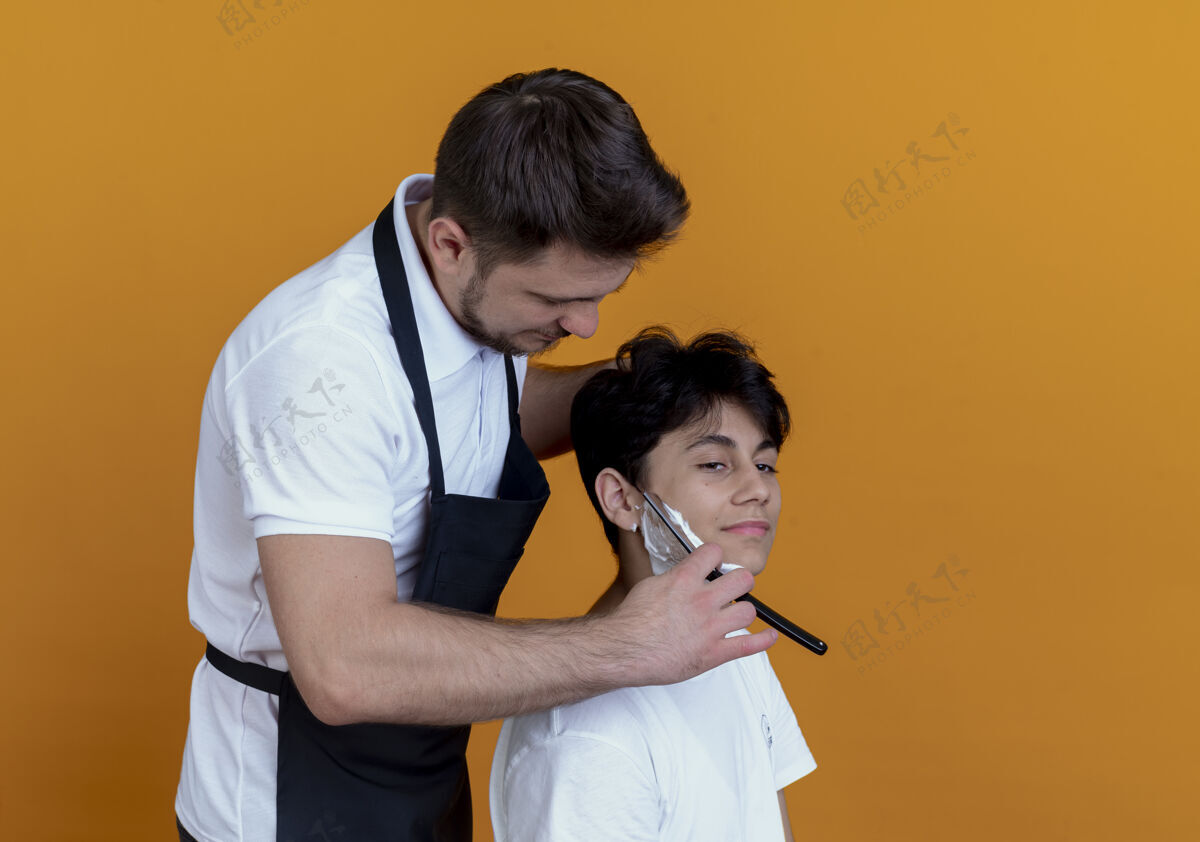 顾客穿着围裙的理发师用满意的顾客剃须刀在橘色墙上刮胡子围裙胡须理发师