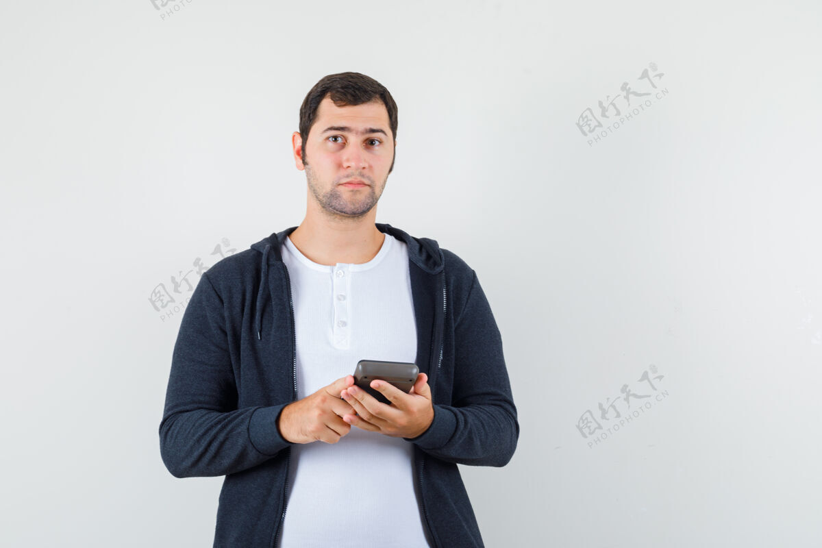 男人年轻人拿着计算器在白色t恤和拉链前黑色连帽衫 看起来严肃 正面视图肖像连帽衫T恤