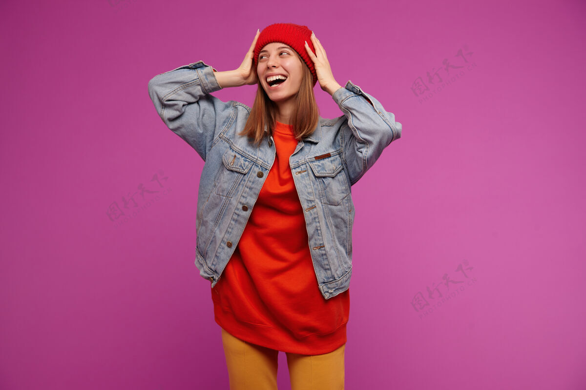 服装一幅迷人的年轻女子的肖像 深色长发穿着牛仔裤夹克 黄色裤子 红色毛衣和帽子在复制空间看左边 隔离在紫色的墙上情感裤子成人