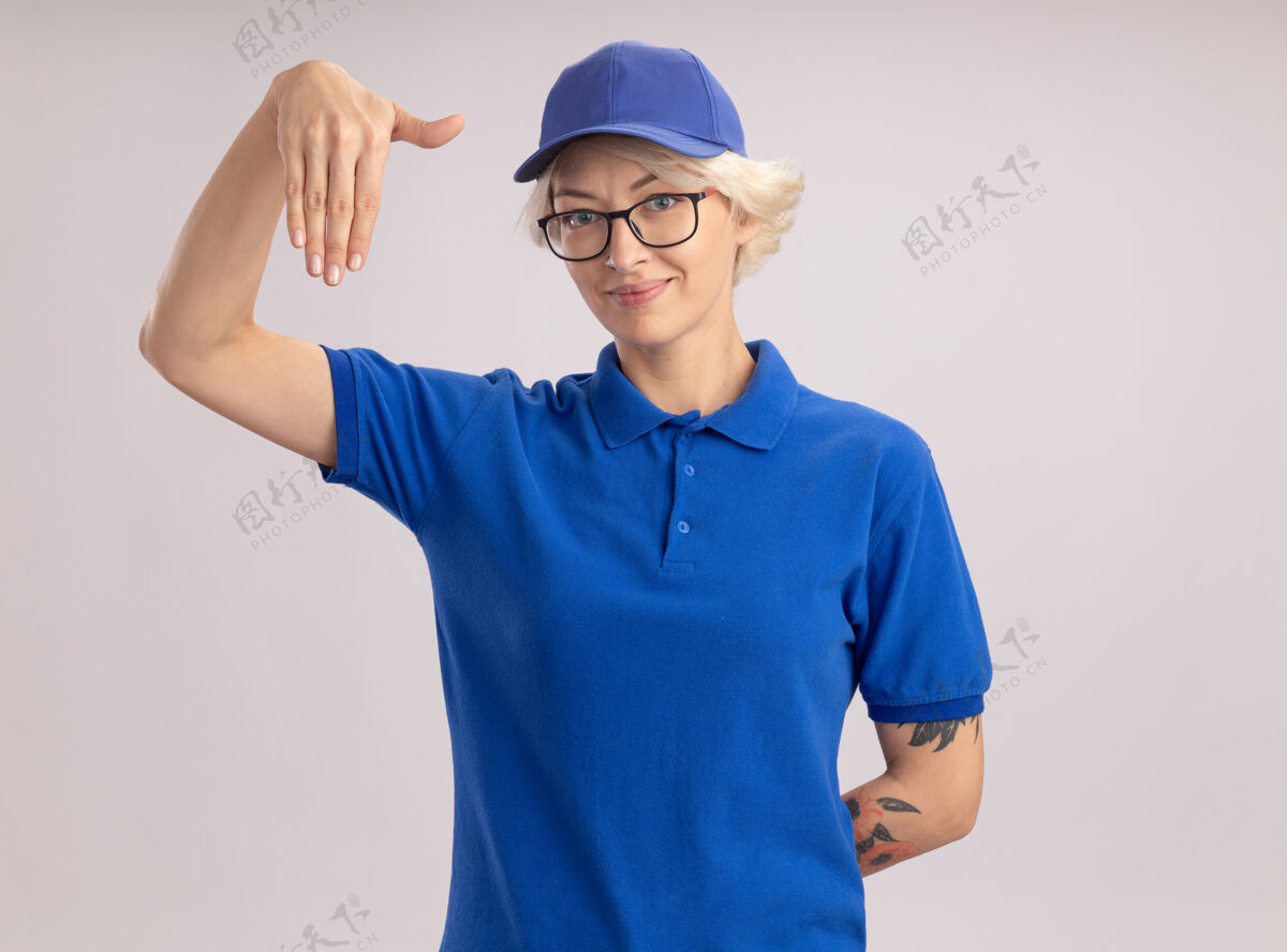 手身穿蓝色制服 戴着眼镜的年轻送货女自信地用手做着手势 站在白色的墙壁上的肢体语言概念帽子信心语言