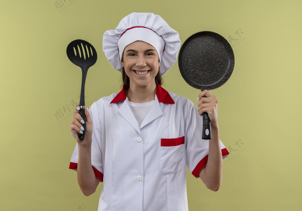 空间身着厨师制服 面带微笑的白人年轻厨师女孩拿着抹刀和煎锅 隔离在有复印空间的绿地上复制锅女孩