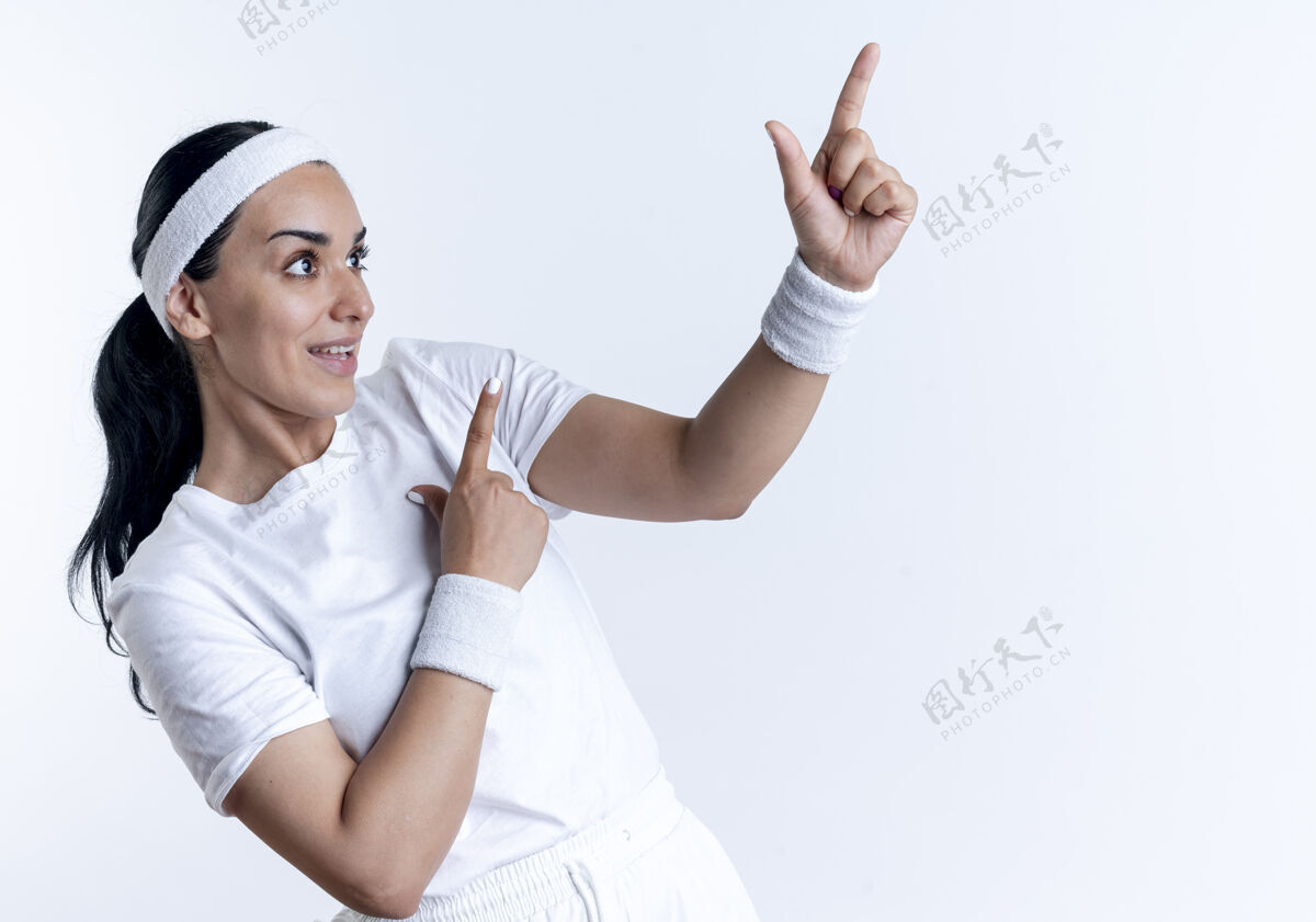 看年轻惊讶的白人运动女性戴着头带和腕带看和点在一边 在白色空间与复制空间隔离壁板头带女人