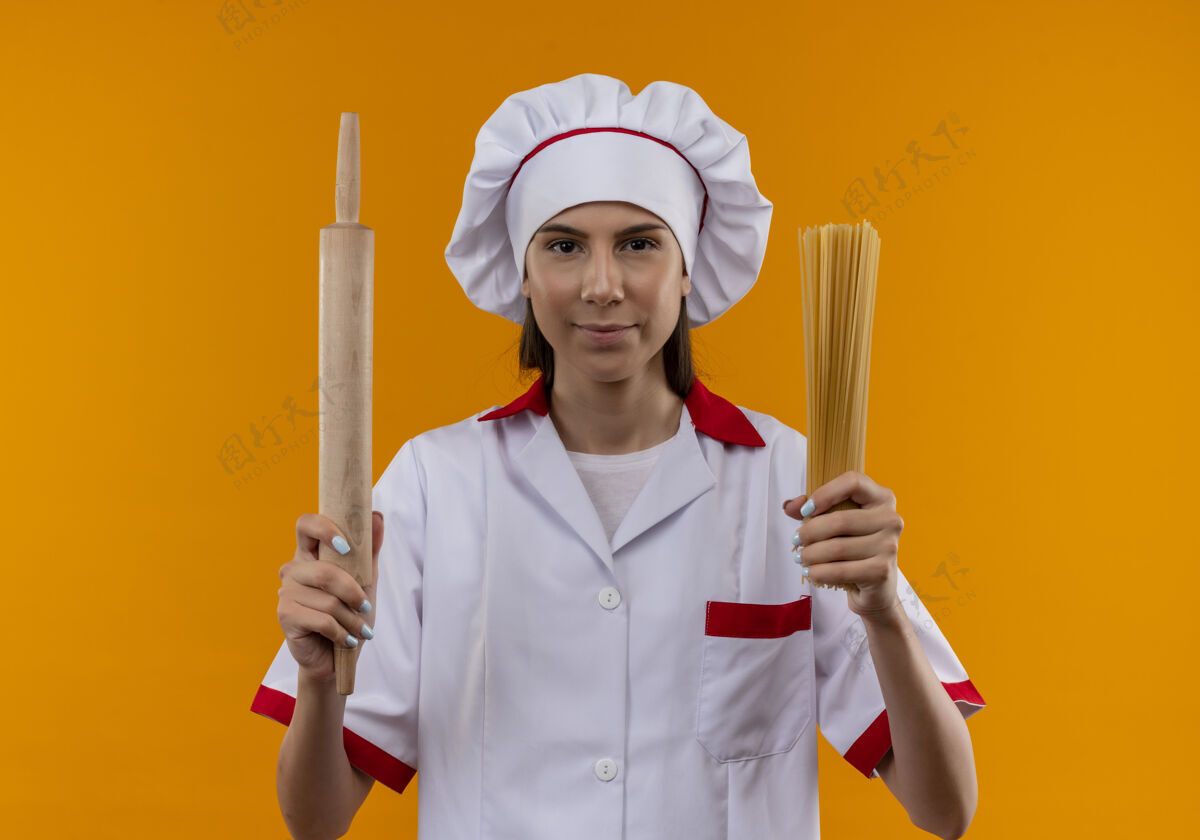 别针身着厨师制服的年轻自信的白人厨师女孩拿着擀面杖和一串意大利面 在橙色的空间里与复制空间隔离开来束年轻复制