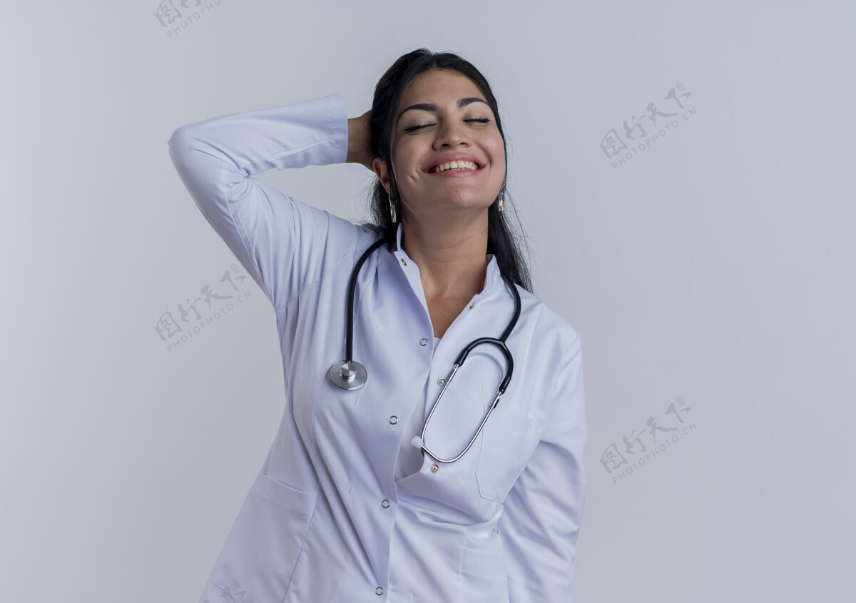 穿面带微笑的年轻女医生 穿着医用长袍 手持听诊器 将手放在脑后 闭上眼睛 隔离在白色墙壁上 留有复印空间手微笑年轻