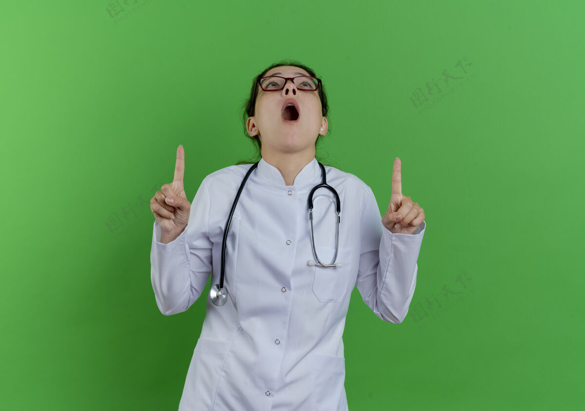 穿年轻的女医生穿着医用长袍 戴着听诊器 戴着眼镜 在绿色的墙上看着并指着隔离的复制空间指点女听诊器