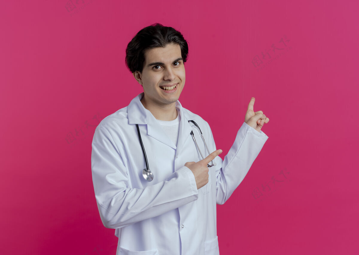 年轻面带微笑的年轻男医生穿着医用长袍 听诊器指着身后隔离的粉红色墙上有复印空间穿指着看