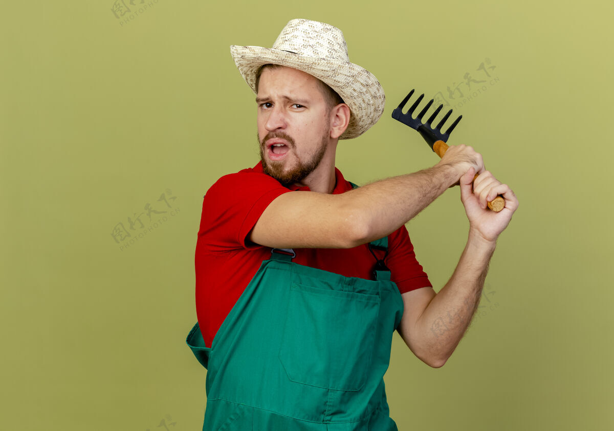 持有年轻英俊的斯拉夫园丁 穿着制服 戴着帽子 拿着耙子 准备用耙子打一个人 耙子被隔离在橄榄绿的墙上 留着复制空间耙花园年轻