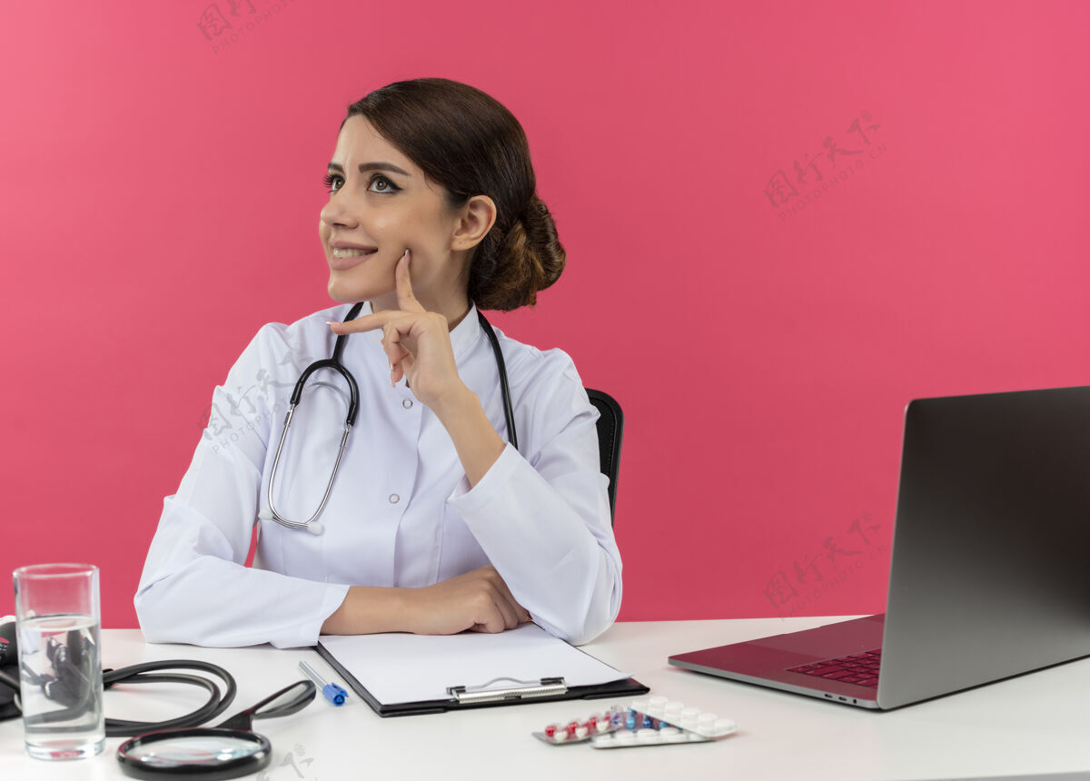 微笑面带微笑的年轻女医生穿着医用长袍和听诊器坐在办公桌旁 手里拿着医疗工具和笔记本电脑 把头转向一边 抬起头抚摸脸颊 手指被隔离在粉红色的墙上穿听诊器壁板