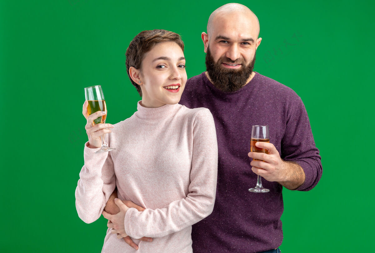 一起一对穿着休闲服的快乐年轻夫妇一个留着短头发和胡须的女人一个戴着香槟的男人一起来欢庆情人节站在绿色的墙上情侣衣服女人