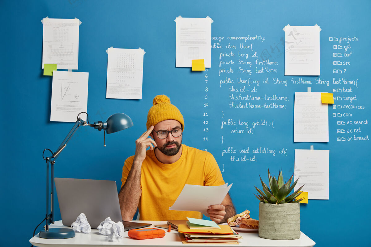 工人严肃的男性员工或自由职业者考虑纸质文件 戴黄色帽子和t恤 在线学习笔记本电脑 在家工作 浏览资料 在同事的空间里摆姿势计划男性工作