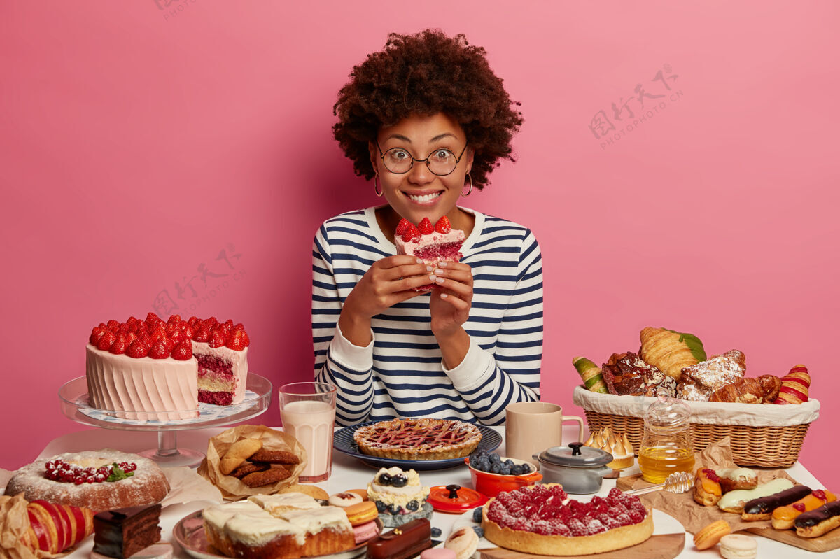 室内快乐的黑皮肤女人吃着美味的草莓蛋糕 穿着条纹套头衫 在餐桌上摆满了甜点 得到了极大的乐趣 在玫瑰色的墙上摆姿势煎饼甜点桌子