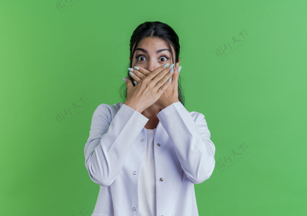 穿惊讶的年轻女医生穿着医用长袍捂着嘴 双手隔离在绿色的墙上 留着复印空间女性手长袍