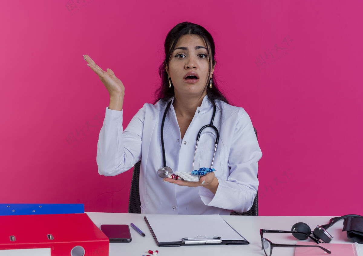 困惑困惑的年轻女医生穿着医用长袍和听诊器坐在办公桌旁 手里拿着医疗工具 手里拿着药品 粉红色的墙上空手孤立着医疗看年轻