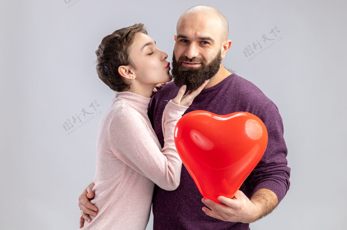 年轻一对穿着休闲服的年轻情侣 幸福的女人用心形气球亲吻留胡子的男朋友 站在白色背景上庆祝情人节胡须庆祝吻