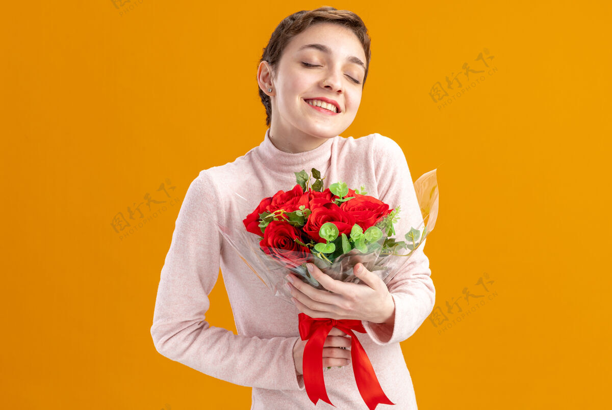 女人一个留着短发的年轻女子 手里捧着一束红玫瑰 快乐而乐观 闭着眼睛 微笑着站在橘色的墙上庆祝情人节微笑年轻立场