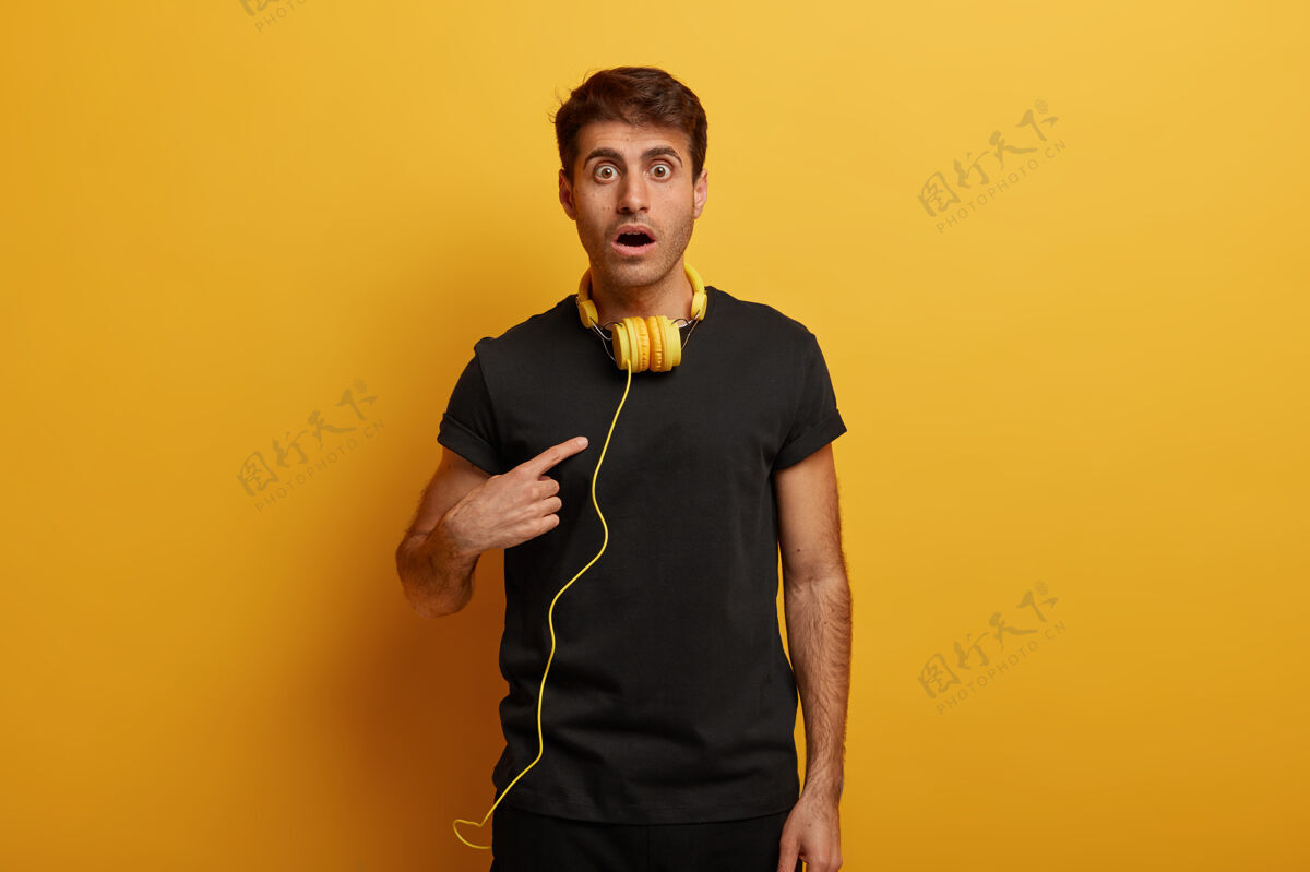 人类白种人指着自己 觉得被选中很震惊 张大嘴巴 用耳机上网听广播 隔离在黄色的墙上不高兴休闲男性