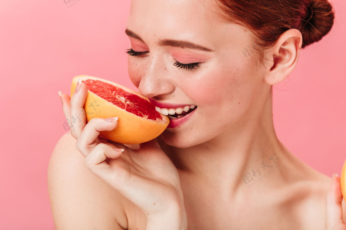 脸快乐的年轻女人在吃葡萄柚微笑的姜女士在粉红色的背景下享受柑橘女孩健康一个人