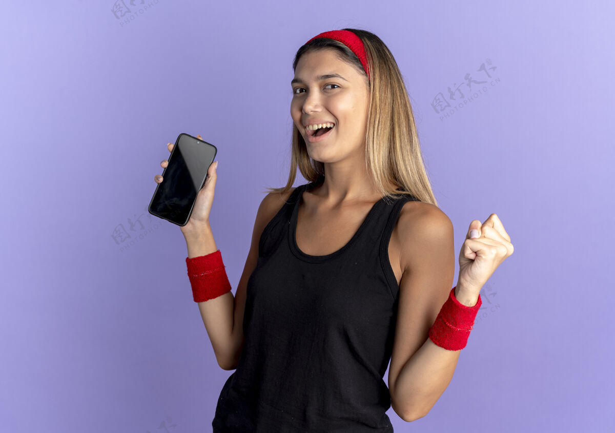 拳头身穿黑色运动服 头戴红色头带 手持智能手机 握紧拳头的年轻健身女孩 为蓝色而高兴和兴奋握紧黑色运动装