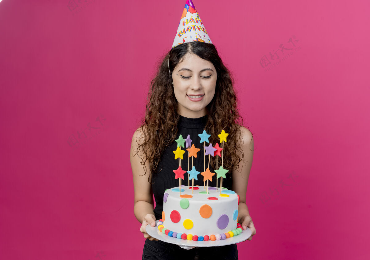 女性一个年轻漂亮的女人 卷发 戴着节日帽 手里拿着生日蛋糕 生日快乐 积极的生日派对概念 粉色的蛋糕派对快乐