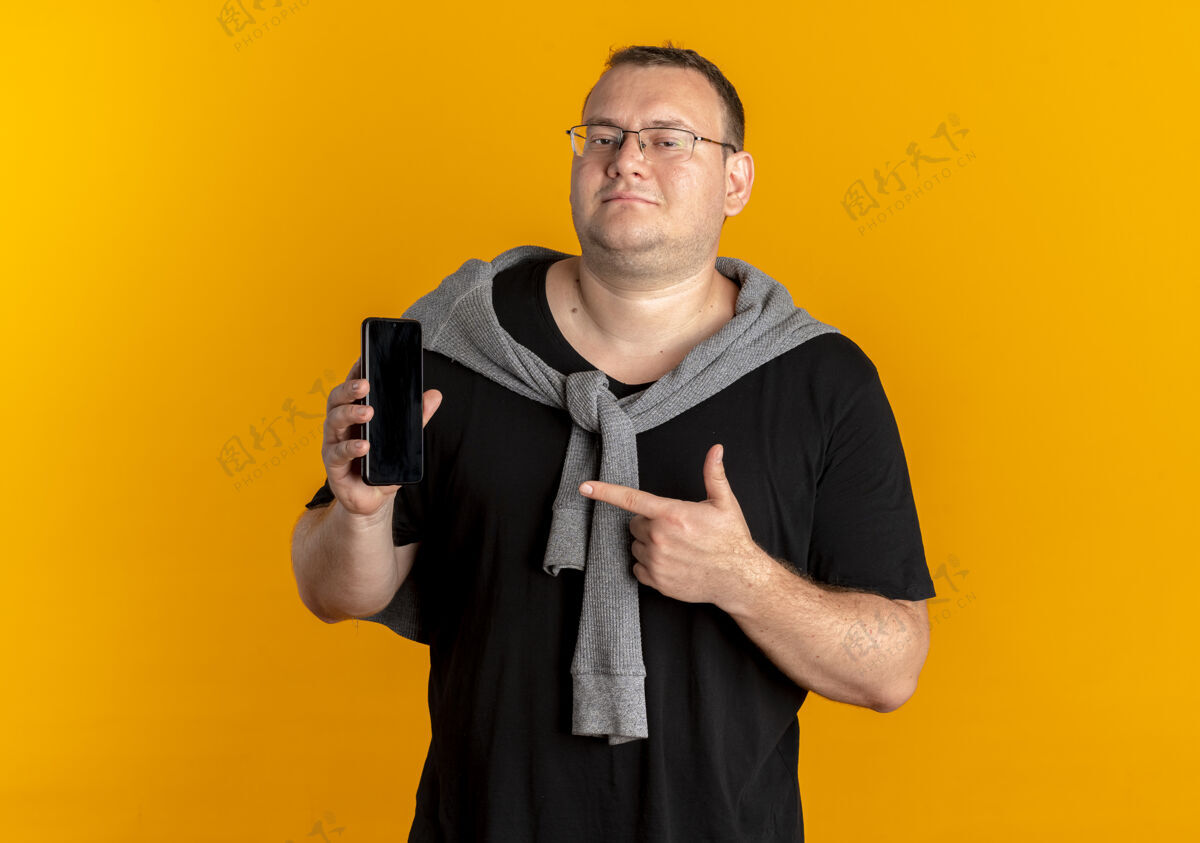 手指戴眼镜的超重男子身穿黑色t恤 展示智能手机 手指指着它 脸上露出橙色的微笑站着黑色T恤