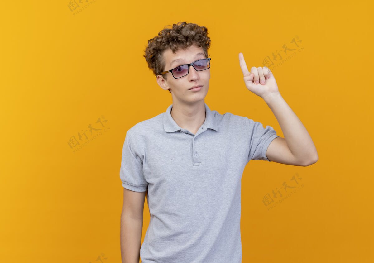 看戴着黑眼镜的年轻人穿着灰色马球衫 高兴而积极地展示食指对橙色有新的想法站创意男人