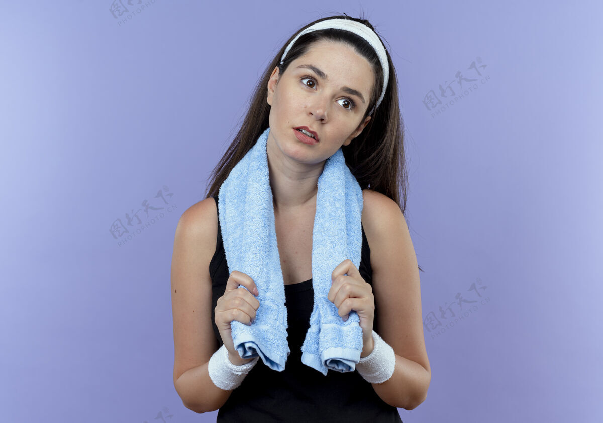 年轻戴着头巾脖子上围着毛巾的年轻健身女士站在蓝色的墙上 一边看一边又累又无聊健身女人站