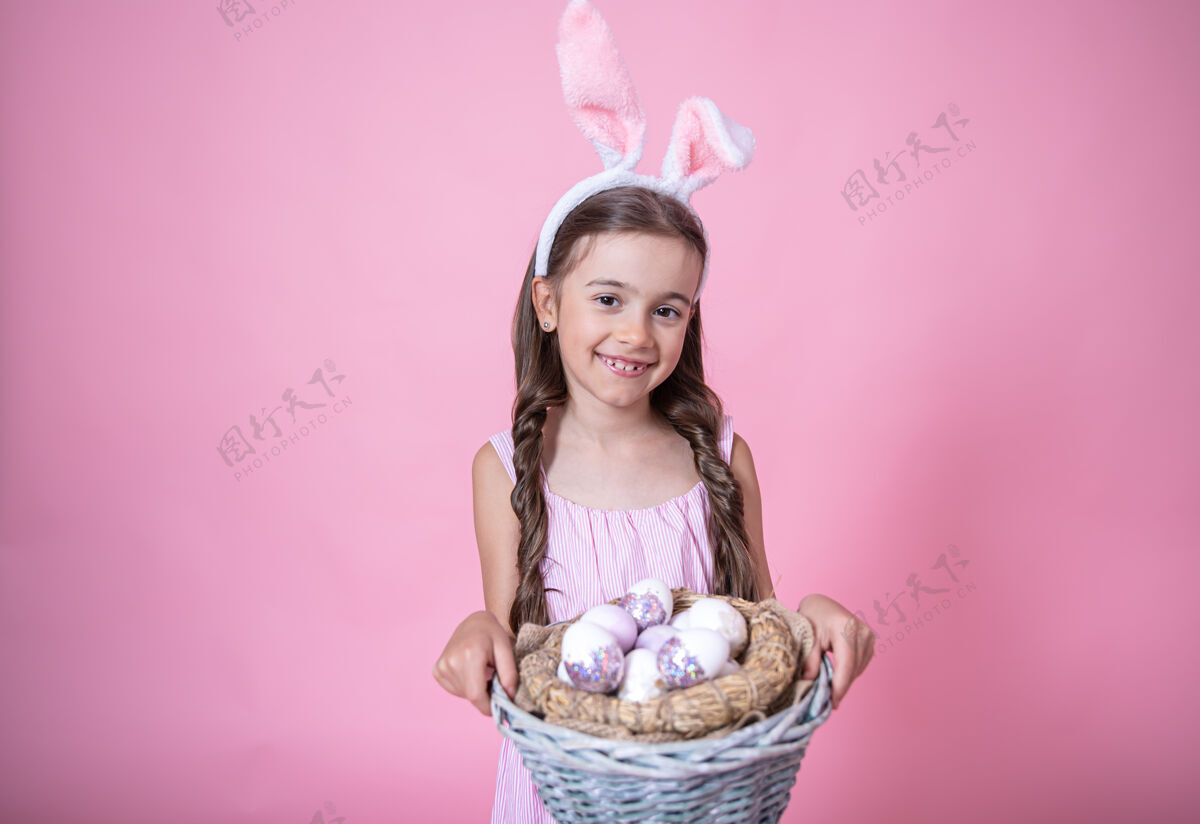 篮子在粉红色的工作室里 一个小女孩拿着一个装满复活节彩蛋的篮子摆着复活节兔子耳朵的姿势兔子节日孩子们