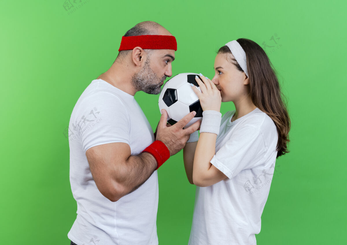 看一对戴着头带和腕带的成年运动情侣 手拿着足球 亲吻着足球 看着对方抱着腕带球