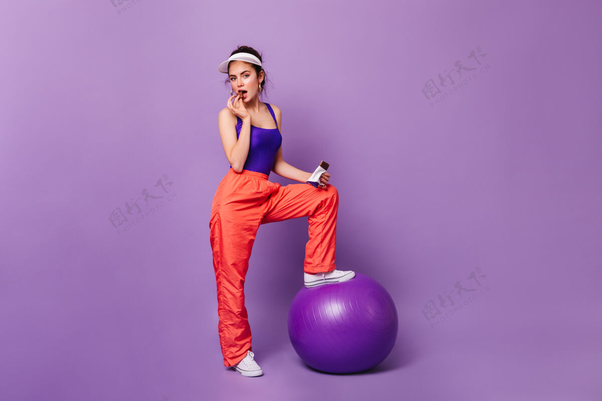 成人穿着运动装的调情女人和fitball在紫色墙上摆姿势运动橙色裤子头