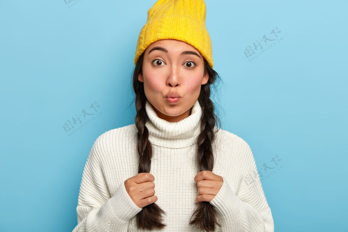 套头衫迷人的女人嘴唇圆润 留着两条辫子 穿着白色保暖毛衣 戴着黄色帽子 在镜头前有着挑逗的神情 隔着蓝色的墙壁头像冬天亚洲人
