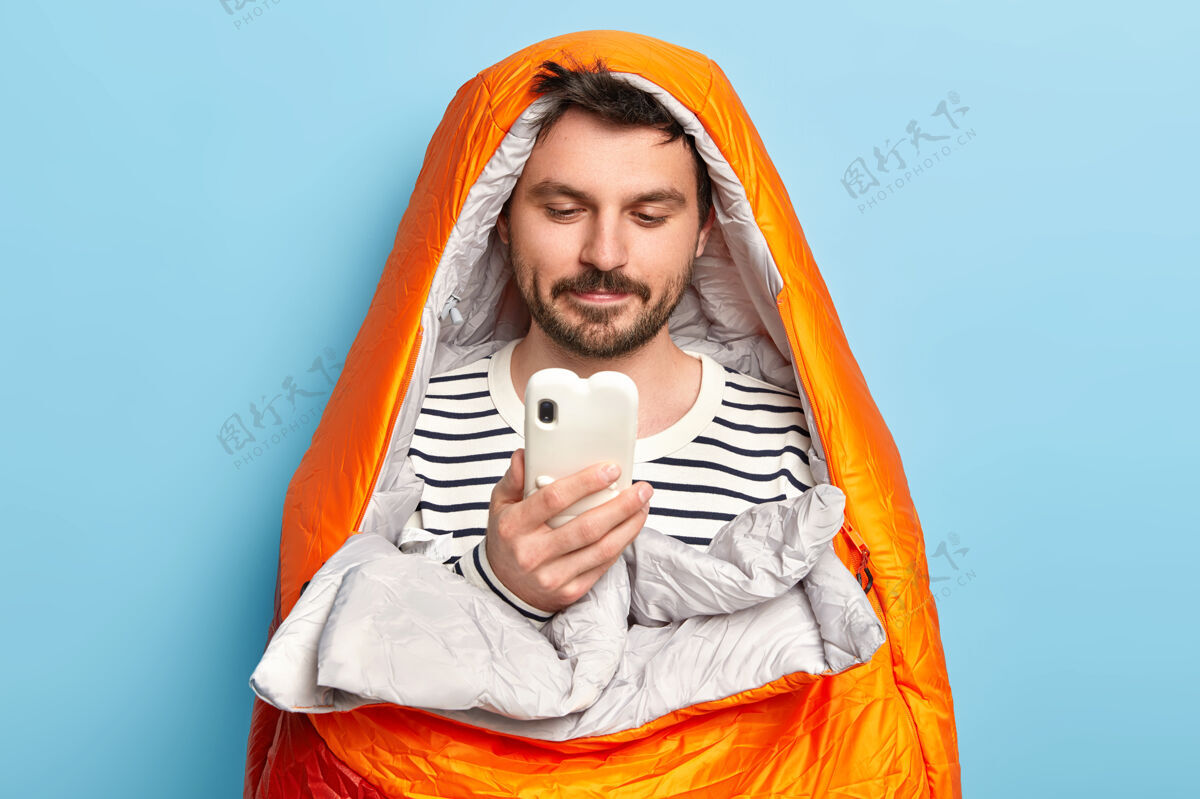人男性旅行者在橙色温暖的睡袋里摆姿势 在海边度过休闲时光 专注于智能手机 在室内找到合适的目的地摆姿势旅行人活动