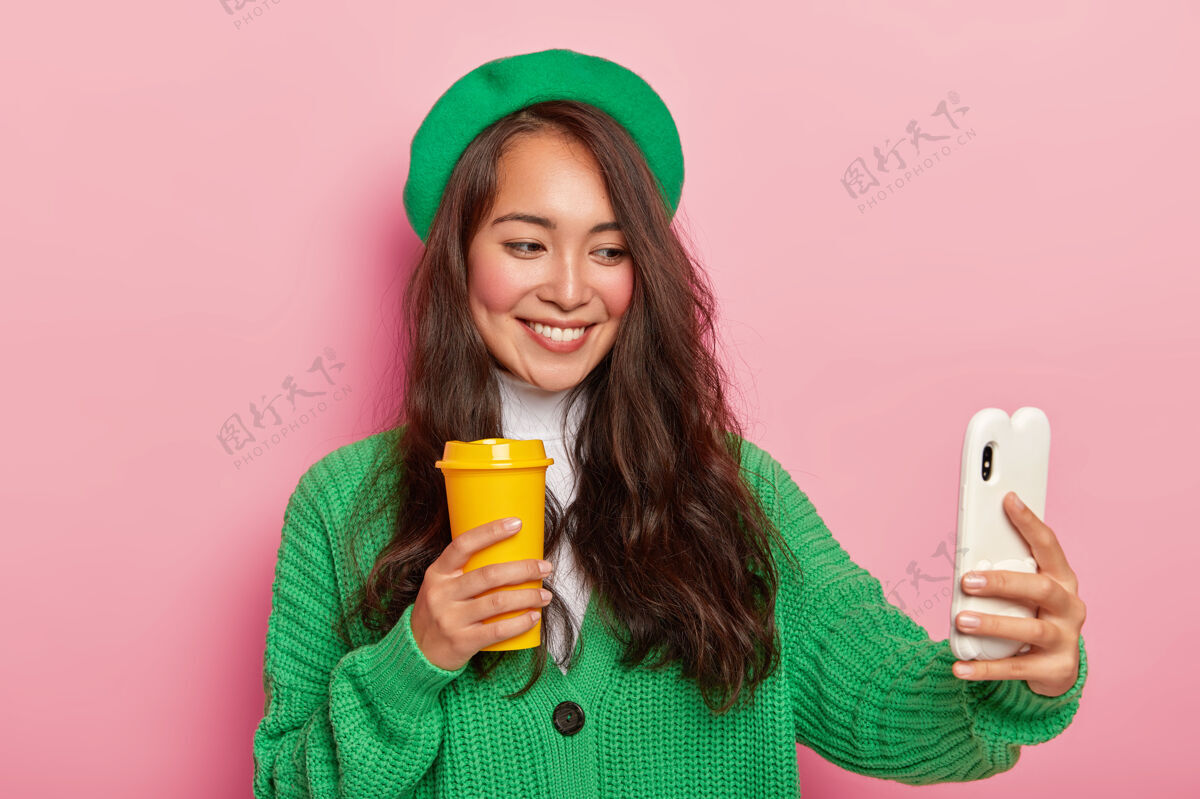杯子身着绿色贝雷帽和针织毛衣的快乐混血女士 用手机自拍肖像 用咖啡杯摆姿势休闲请表情