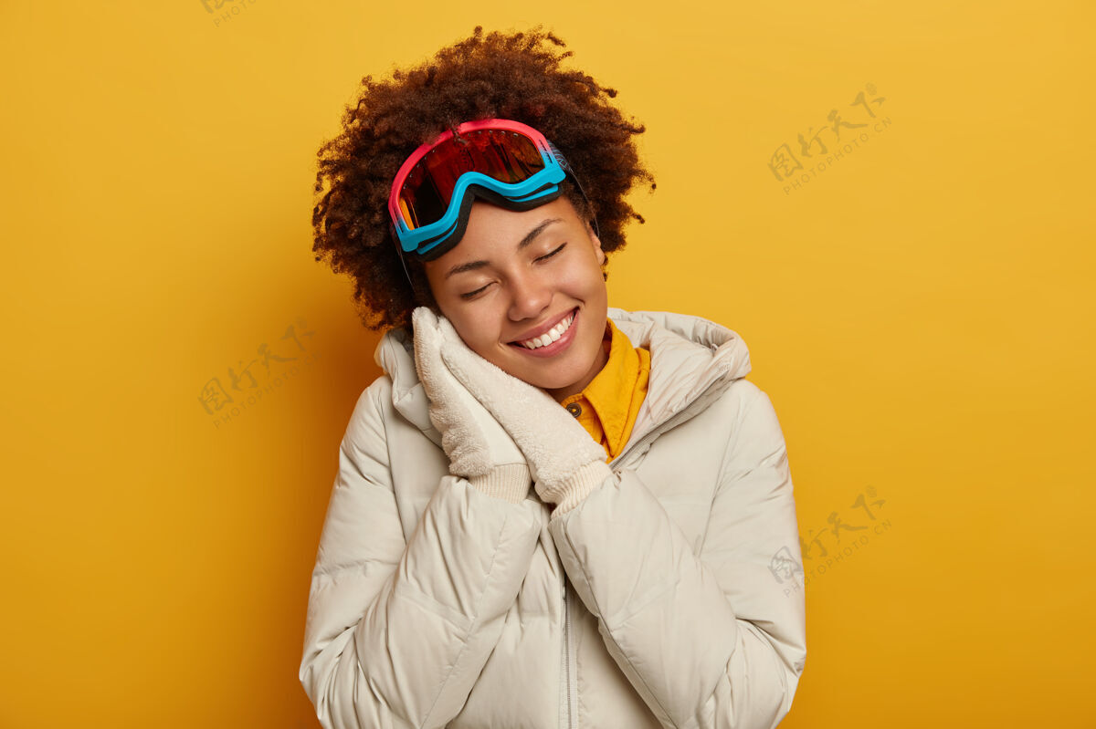 温柔非洲式发型的积极女性 斜靠在紧握的手掌上 有着梦幻般的面部表情 穿着白外套 戴着手套和滑雪面罩 享受冬天的冒险 隔离在黄色的墙上快乐女性冬季