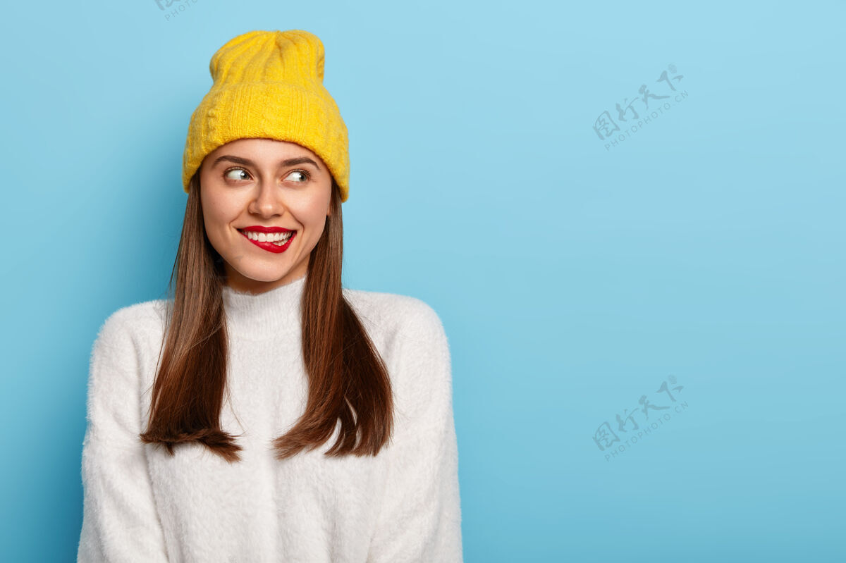 肖像迷人的年轻女子咬着红唇 想着什么 转眼望去 乌黑的直发 戴着黄色的帽子 穿着暖和的白色毛衣温柔高兴帽子